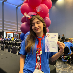 Fabiola González at DI's Global Finals 2024 event. She is wearing a blue shirt and a DI alumni red tie with various pins attached to it. Fabiola is holding up her Champion of Inclusion Award certificate. 