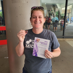 Tonya Hernández is wearing a gray DI Michigan t-shirt and is smiling. She is holding her Champion of Inclusion pin and certificate. 